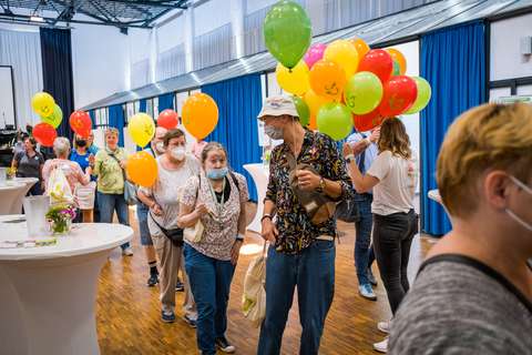 Mehrere Personen stehen in einem Raum mit Luftballons in den Händen, einige tragen Masken.