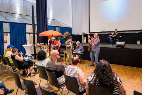Mehrere Personen sitzen auf Stühlen in einem Saal, während vorne einige Personen musizieren und eine Person singt.