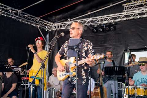 Mehrere Personen musizieren gemeinsam auf einer Bühne, eine Person spielt E-Gitarre und singt, andere spielen verschiedene Instrumente.