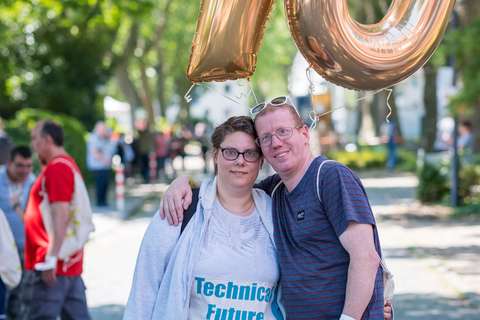 Zwei Personen stehen eng umarmt im Freien, über ihnen goldene Luftballons in Form der Zahl 70.