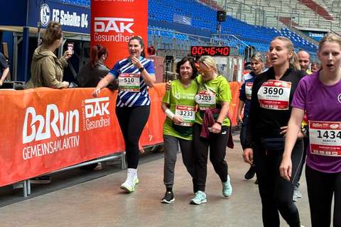 Mehrere Personen mit Startnummern gehen oder laufen in einer Halle an einer Absperrung mit B2Run-Logo vorbei, im Hintergrund sind Tribünen und eine große Uhr sichtbar.