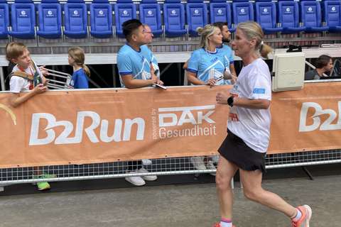 Eine Person läuft an einer Absperrung mit B2Run-Banner vorbei, dahinter stehen mehrere Personen vor leeren blauen Stadion-Sitzen.