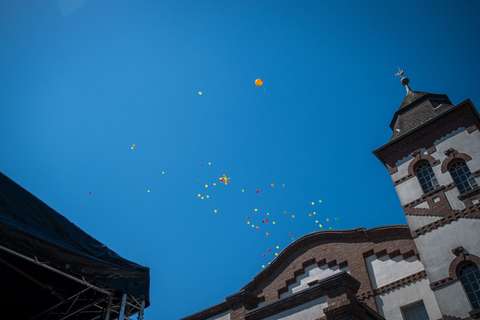 Mehrere bunte Luftballons steigen in den blauen Himmel, rechts ein historisches Gebäude mit Turm, links ein Teil einer Bühnenkonstruktion.