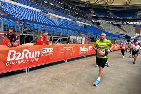 Mehrere Personen laufen in einem Stadion an einer Absperrung mit B2Run-Werbung vorbei, während zwei Personen in orangefarbener Kleidung daneben stehen.