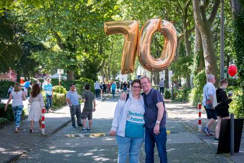 Zwei Personen stehen eng beieinander auf einer Straße und halten goldene Luftballons in Form der Zahl 70, im Hintergrund sind weitere Personen und Bäume.