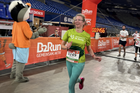 Eine Person läuft mit Startnummer 3249 durch einen Zielbogen bei einer Laufveranstaltung in einem Stadion, während eine weitere Person im Maskottchenkostüm am Rand steht.