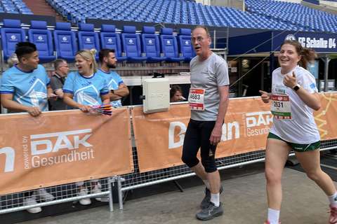 Zwei Personen laufen in einem Stadion an einer Absperrung mit orangefarbenem Banner vorbei, im Hintergrund stehen mehrere Personen in blauen T-Shirts.