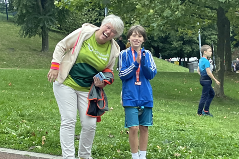 Zwei Personen stehen auf einer Wiese vor Bäumen, eine Person hält Kleidung in der Hand, im Hintergrund läuft ein Kind.