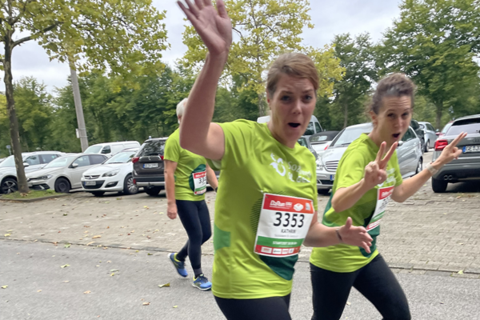 Drei Personen in grünen T-Shirts mit Startnummern joggen auf einer Straße, zwei davon winken und zeigen Gesten in die Kamera.