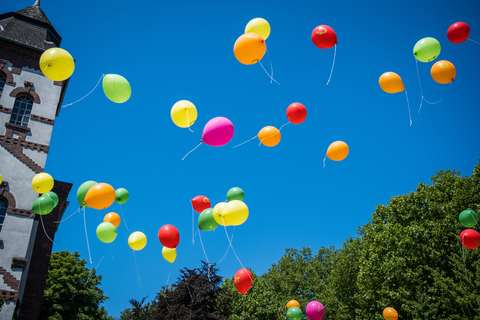 Mehrere bunte Luftballons steigen in den Himmel, im Hintergrund sind Bäume und ein Gebäudeturm.