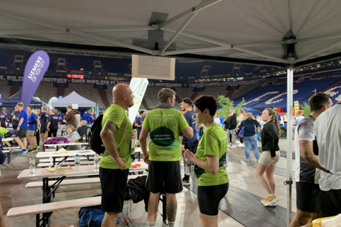 Mehrere Personen in Sportkleidung stehen und sitzen unter einem Pavillon auf einer Stadiontribüne, im Hintergrund weitere Menschen und Bänke.
