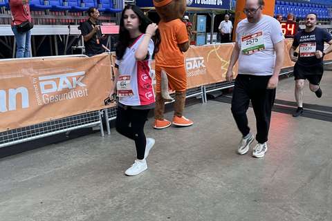 Mehrere Personen gehen oder laufen in einem Stadion an einem orangefarbenen Zielbogen vorbei, im Hintergrund steht eine Person im Tierkostüm.