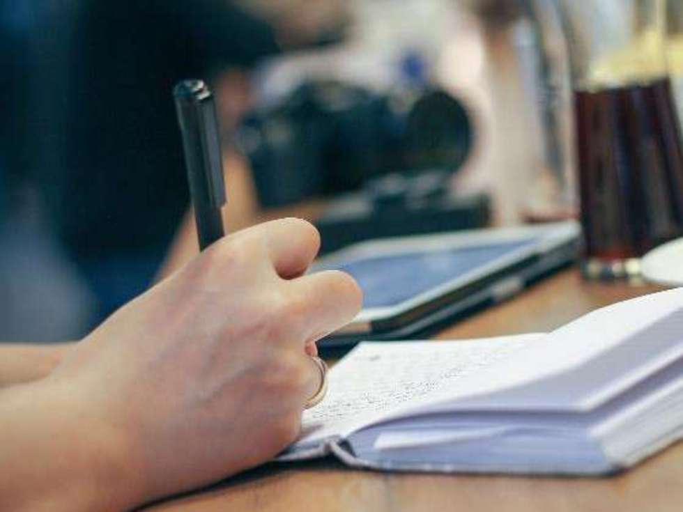Eine Person schreibt mit einem Stift in ein Notizbuch, daneben liegen ein Tablet und ein Glas Getränk.