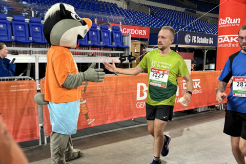 Eine Person im grünen Laufshirt gibt einem Maskottchen in Tierkostüm und orangefarbenem T-Shirt einen High-Five in einer Sportarena.