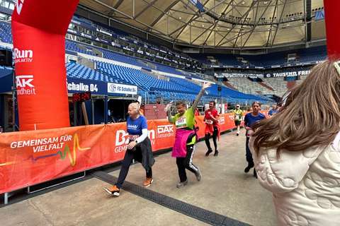 Mehrere Personen gehen oder laufen durch einen Zielbogen in einem Stadion, einige tragen Startnummern und Sportkleidung.