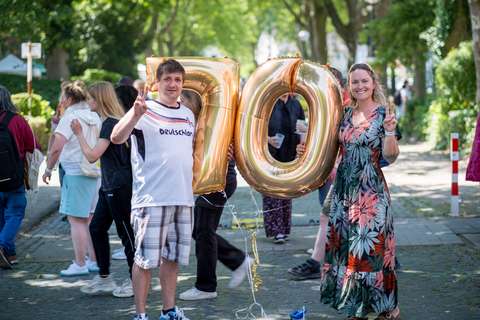 Zwei Personen halten goldene Luftballons in Form der Zahl 70 und zeigen das Victory-Zeichen, im Hintergrund gehen weitere Personen auf einer Straße.