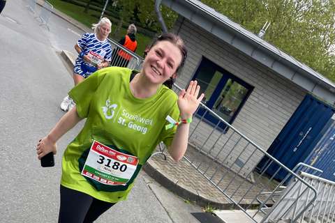Eine Person mit grüner Laufshirt und Startnummer läuft und winkt, im Hintergrund läuft eine weitere Person.