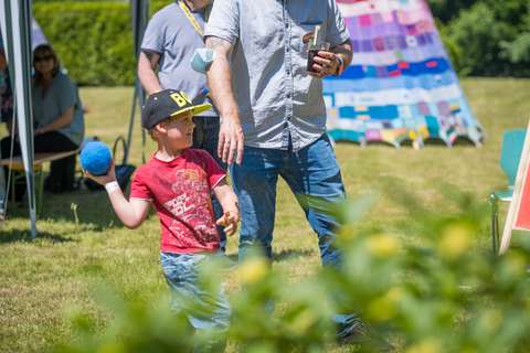 Ein Kind mit schwarzer Kappe und rotem T-Shirt hält einen blauen Ball, daneben steht eine Person mit Getränk in der Hand.