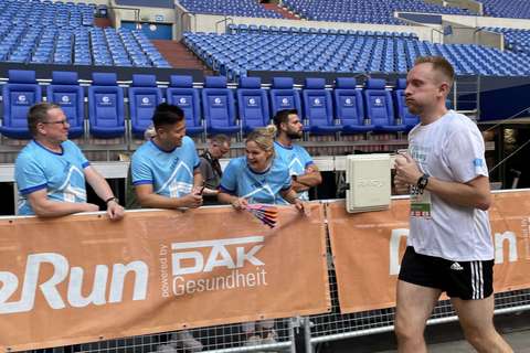 Eine Person läuft an einer Absperrung vorbei, hinter der vier Personen in blauen T-Shirts stehen und applaudieren.