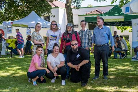 Eine Gruppe von acht Personen posiert im Freien auf einer Wiese, im Hintergrund stehen Pavillons und weitere Personen.