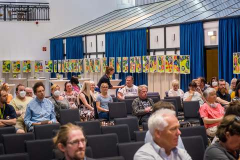 Mehrere Personen sitzen in einem Saal auf Stühlen, einige tragen Masken, im Hintergrund hängen bunte Plakate an einer Wand mit blauen Vorhängen.