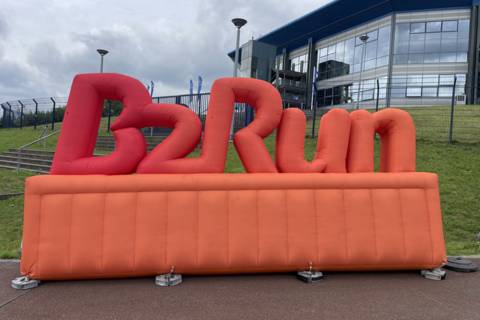 Große, aufblasbare, orangefarbene Buchstaben mit der Aufschrift „B2Run“ stehen vor einem modernen Gebäude und einem Zaun.