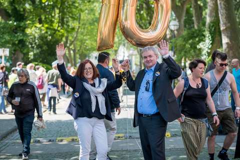 Zwei Personen halten goldene Luftballons in Form der Zahl 10 und heben die Hände, umgeben von weiteren Personen auf einer Straße im Freien.