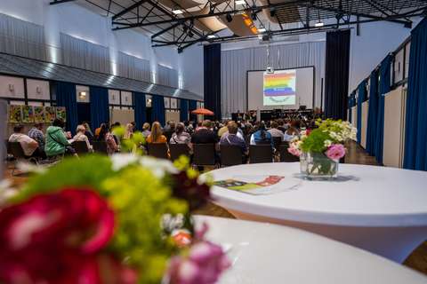 Mehrere Personen sitzen in einem großen Saal und schauen auf eine Bühne mit Leinwand, im Vordergrund steht ein Tisch mit Blumen.