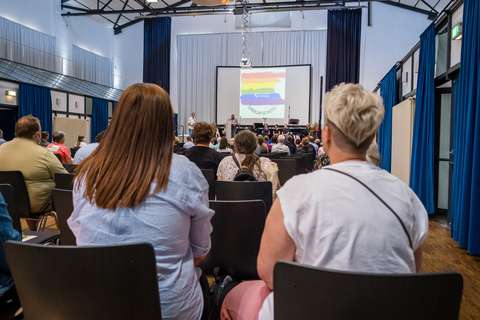 Mehrere Personen sitzen in einem Saal und blicken auf eine Bühne mit einer Leinwand, auf der ein buntes Logo und Text angezeigt werden.
