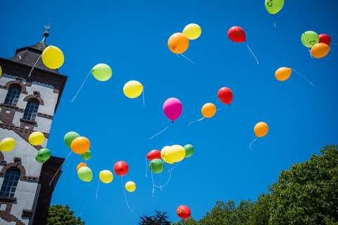 Mehrere bunte Luftballons fliegen vor blauem Himmel, links ein Teil eines Gebäudes und unten Bäume.