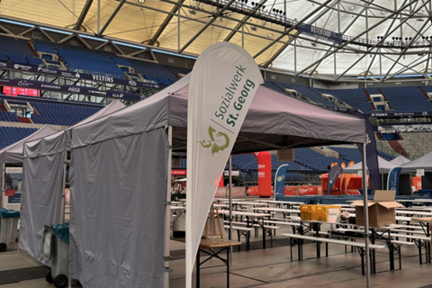 Ein Pavillon mit der Aufschrift „Sozialwerk St. Georg“ steht in einer großen Halle, umgeben von Bierzeltgarnituren und weiteren Ständen.