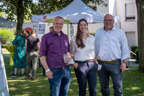 Drei Personen stehen nebeneinander auf einer Wiese vor Pavillons.