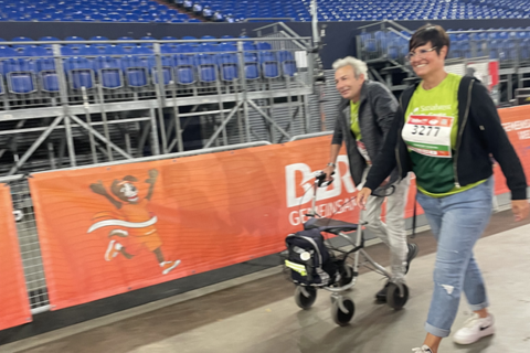 Zwei Personen gehen nebeneinander, eine Person schiebt einen Rollator, im Hintergrund leere Tribünen und ein orangefarbener Zaun mit einer Zeichnung.