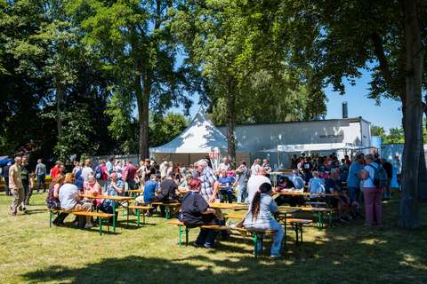 Mehrere Personen sitzen und stehen an Bierzeltgarnituren auf einer Wiese unter Bäumen, im Hintergrund ein weißes Zelt und ein Gebäude.