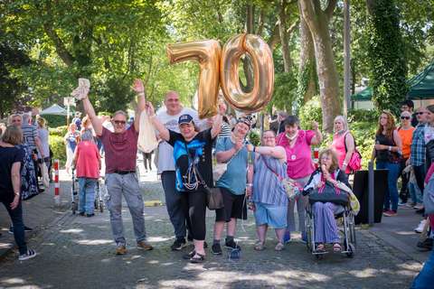 Eine Gruppe von Personen steht auf einer Straße, einige halten goldene Luftballons in Form der Zahl 70, eine Person sitzt im Rollstuhl.