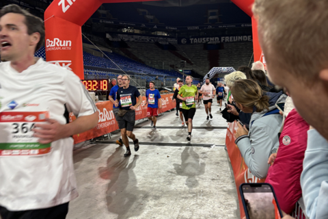 Mehrere Personen laufen durch ein rotes Zieltor bei einer Laufveranstaltung in einem Stadion, während andere am Rand stehen und mit einem Smartphone fotografieren.