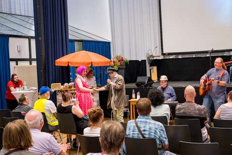 Mehrere Personen befinden sich in einem Saal, einige sitzen im Publikum, während andere vorne musizieren und auftreten; eine Person trägt ein pinkes Kleid und eine pinke Perücke, daneben steht eine Person mit Mikrofon.