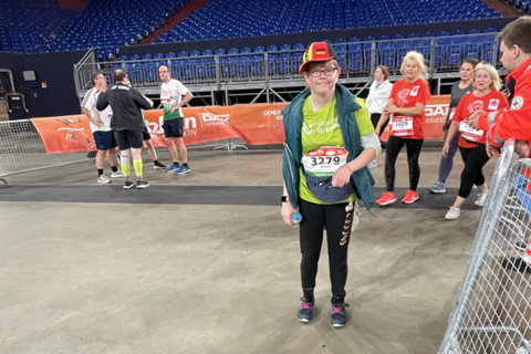 Eine Person mit Startnummer steht in Sportkleidung und bunter Mütze in einer Halle, im Hintergrund mehrere weitere Personen und leere Tribünen.