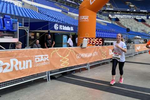 Eine Person läuft unter einem orangefarbenen Zielbogen mit der Aufschrift „B2Run“ und einer digitalen Uhr, im Hintergrund sitzen mehrere Personen auf blauen Tribünenplätzen in einem Stadion.