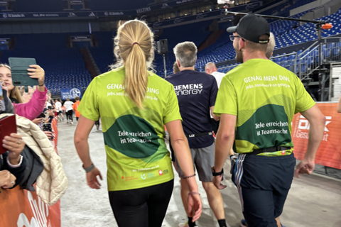 Zwei Personen in Sportkleidung gehen nebeneinander in einer Halle, umgeben von weiteren Personen und Tribünen.