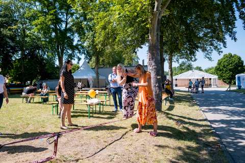 Mehrere Personen stehen und sitzen auf einer Wiese mit Bäumen, Bänken und Zelten; eine Person balanciert auf einem Slackline-Band, während andere zuschauen.