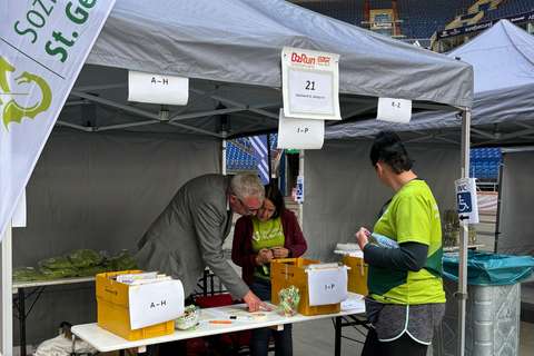 Drei Personen stehen an einem Stand mit mehreren Kisten und beschrifteten Schildern unter einem Pavillon.