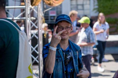 Eine Person mit Brille und Basecap hält zwei Finger vor das Gesicht, im Hintergrund mehrere Personen im Freien.