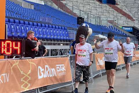 Drei Personen mit Startnummern überqueren eine Ziellinie in einem Stadion, daneben steht eine Person mit Mikrofon und ein Maskottchen.