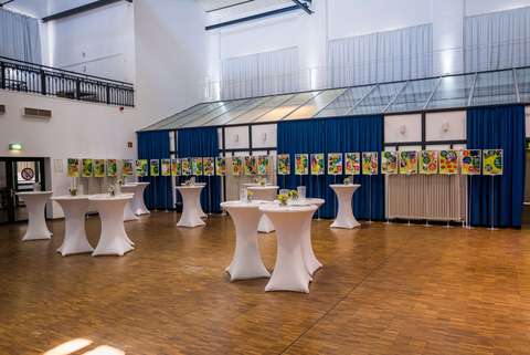 Mehrere Stehtische mit weißen Hussen und kleinen Blumengestecken stehen in einem großen, hellen Raum mit Holzboden; an einer Wand hängen bunte Bilder vor blauen Vorhängen.