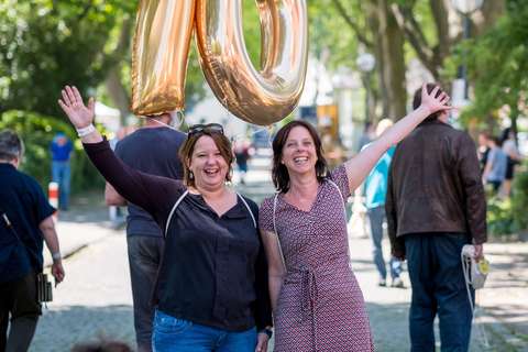 Zwei Personen stehen nebeneinander und halten goldene Luftballons in Form der Zahl 70, im Hintergrund sind weitere Personen und Bäume.