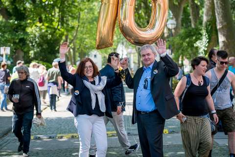 Zwei Personen halten goldene Luftballons in Form der Zahl 10 und heben die Arme, umgeben von weiteren Personen auf einer Straße im Freien.