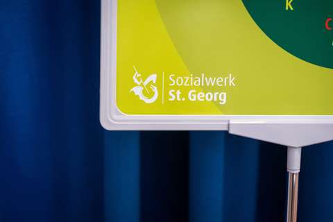Ausschnitt eines Schildes mit der Aufschrift „Sozialwerk St. Georg“ und einem stilisierten Logo auf gelbem Hintergrund.