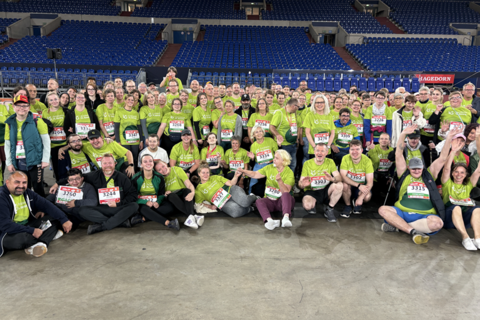 Eine große Gruppe von Personen in grünen T-Shirts mit Startnummern sitzt und steht in einer Halle vor leeren Tribünen.