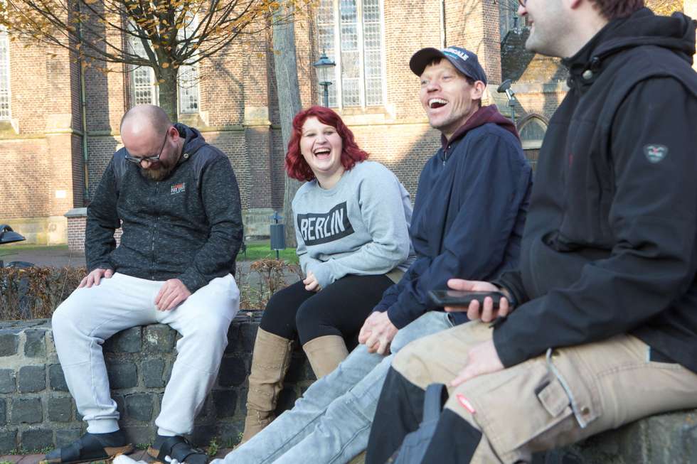 Vier Personen sitzen lachend auf einer niedrigen Mauer im Freien vor einem Gebäude aus Backstein.